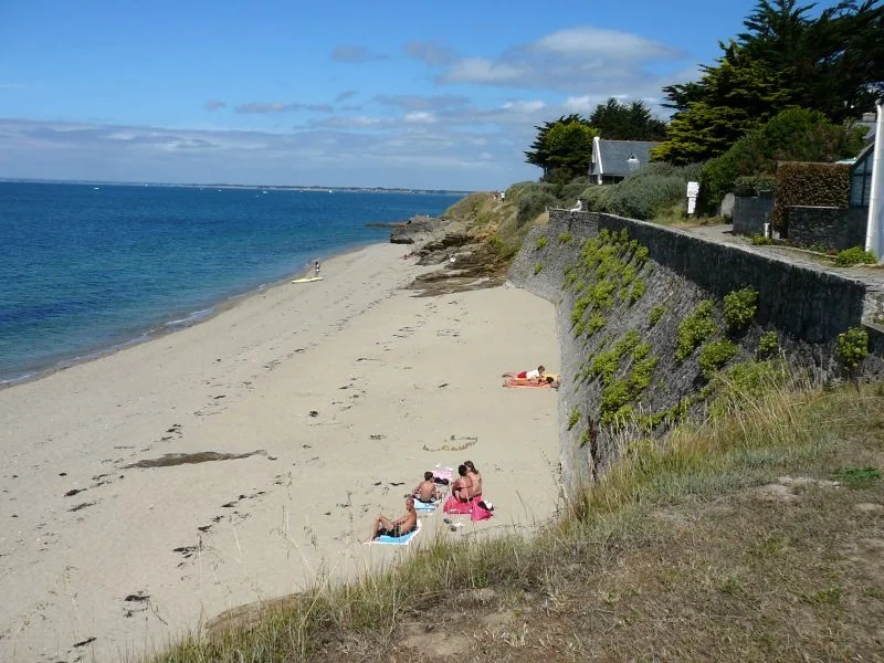 Plage de sable doré à Port-au-Loup avec son mur de soutènement en pierres, baigneurs et vue sur l'océan